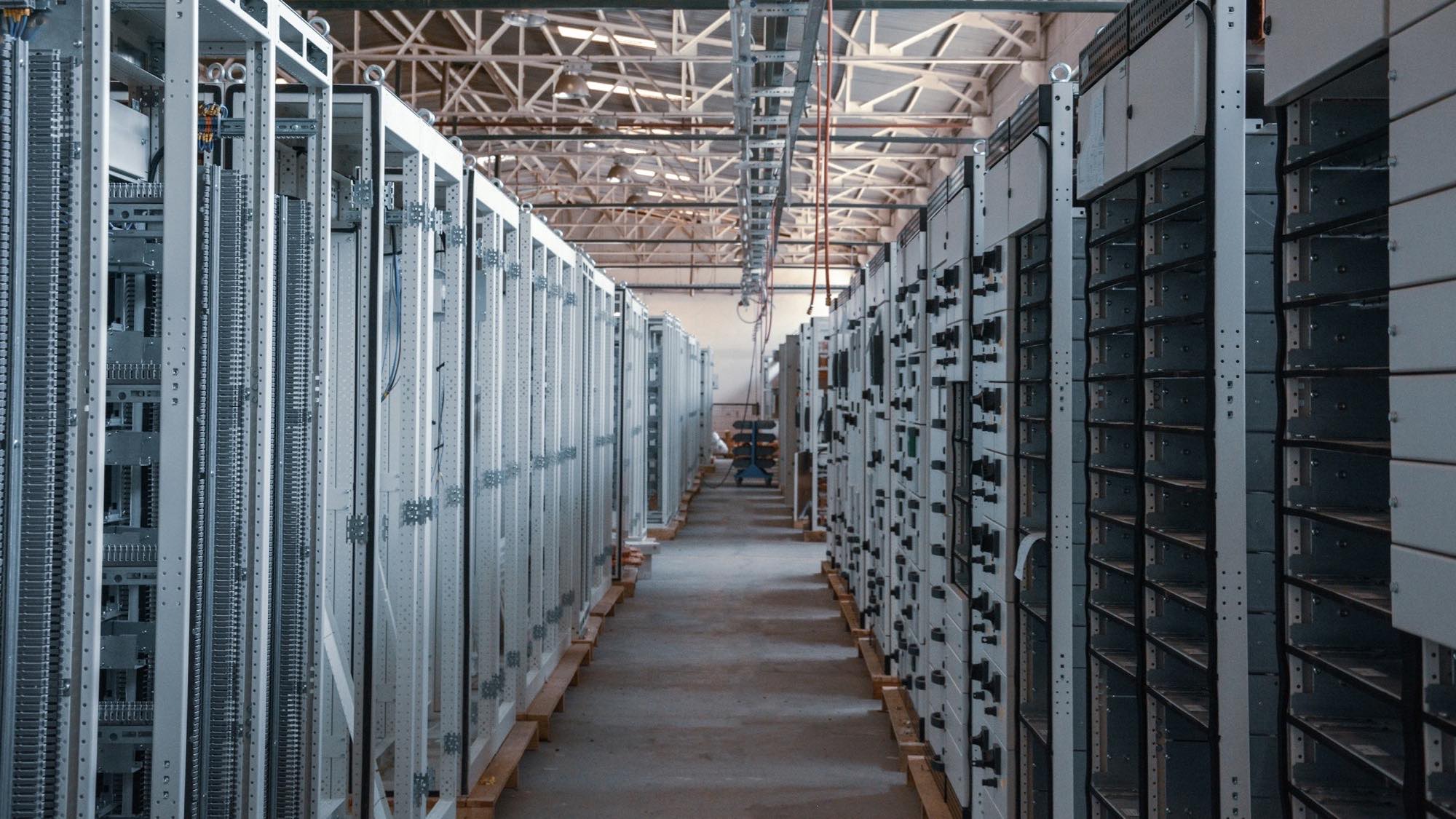 A long aisle receding between two rows of tall metal equipment racks in a data centre, lit from a skylit ceiling
